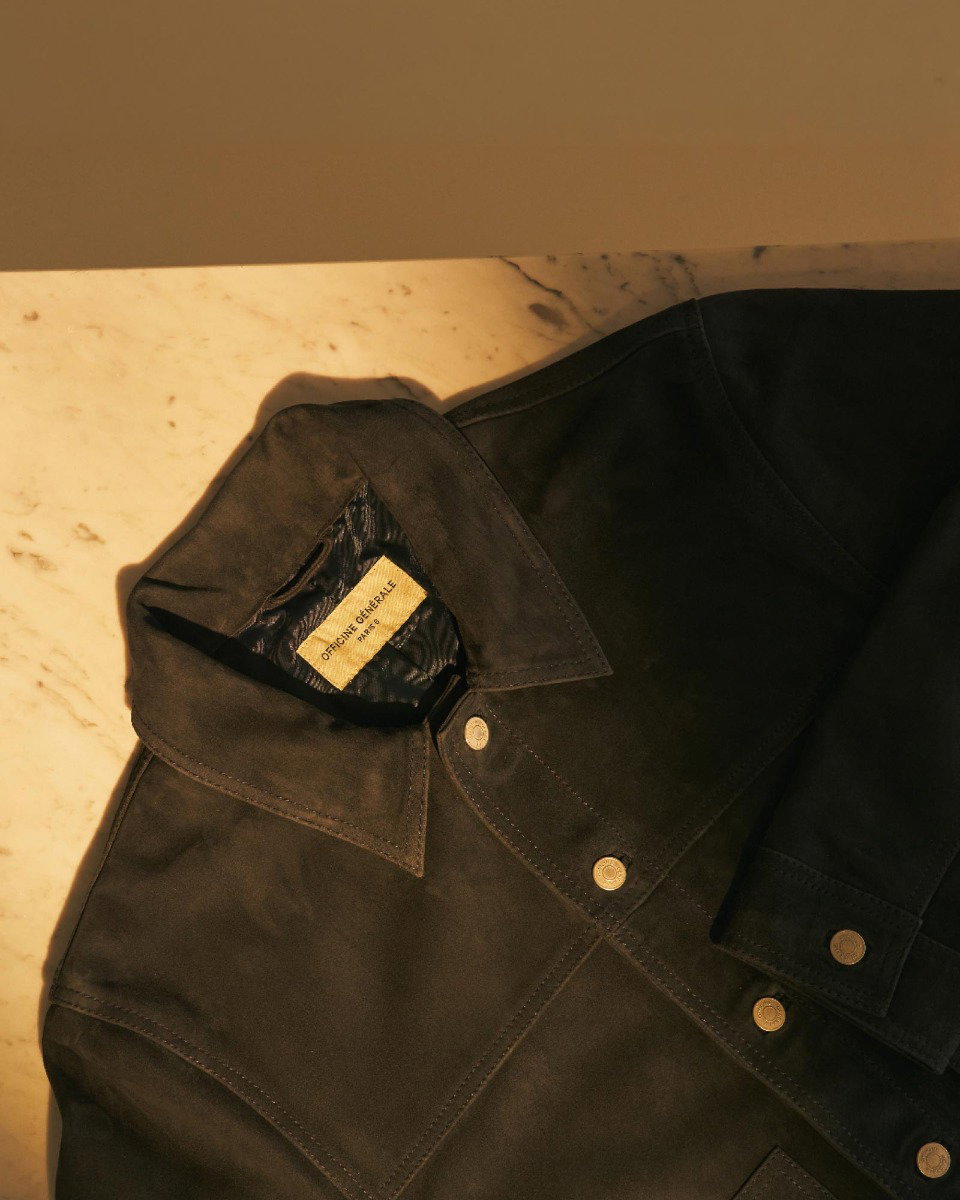 Schwarze Wildlederjacke mit Kragen und silbernen Druckknöpfen, liegend auf marmorierter Oberfläche, Sonnenlicht fällt darauf.