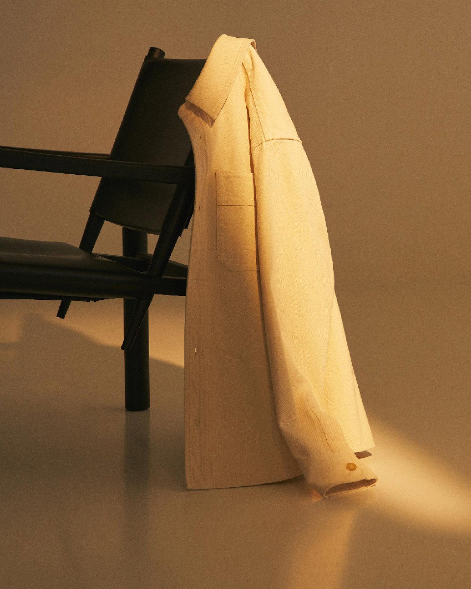 Beige long-sleeve shirt hanging over the back of a black chair with soft warm lighting and a plain background.