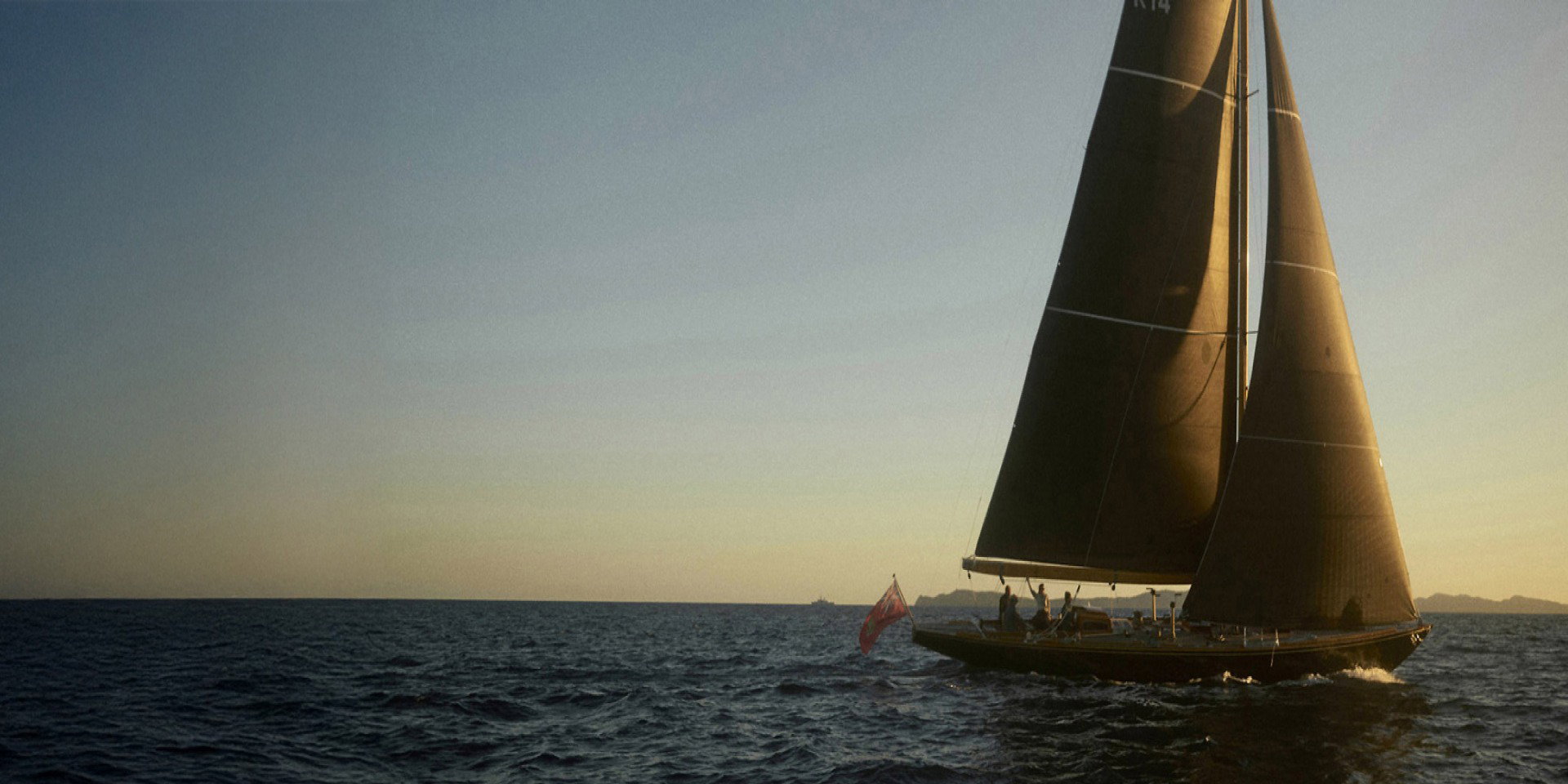 Segelboot mit dunklen Segeln und zwei Personen auf offener See bei Sonnenuntergang, ruhiges Wasser und klarer Himmel.