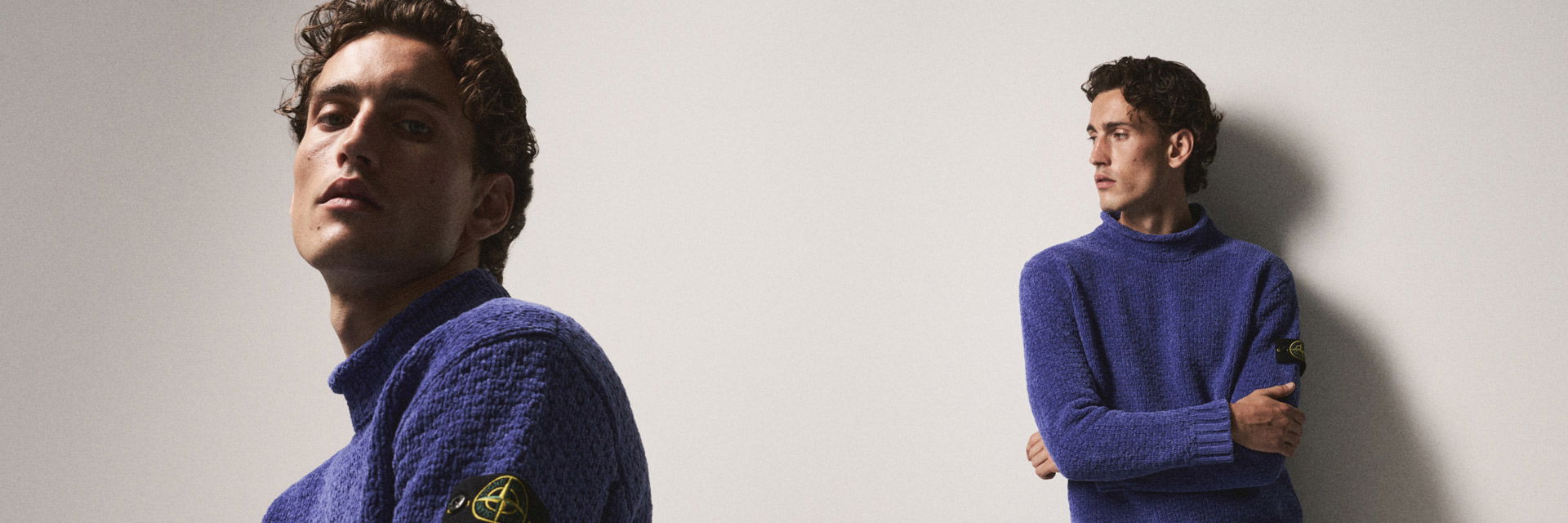 Man with curly hair wearing a textured blue Stone Island sweater, shown in two poses against a plain light wall. Man with curly hair wearing a textured blue Stone Island sweater, shown in two poses against a plain light wall.