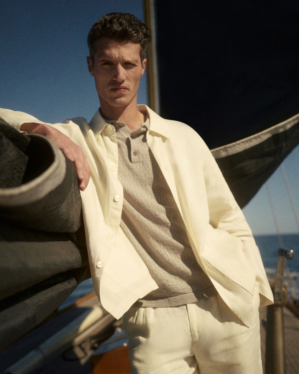 Young man with short curly hair wearing a beige textured polo and cream jacket and pants, leaning on a sailboat railing.