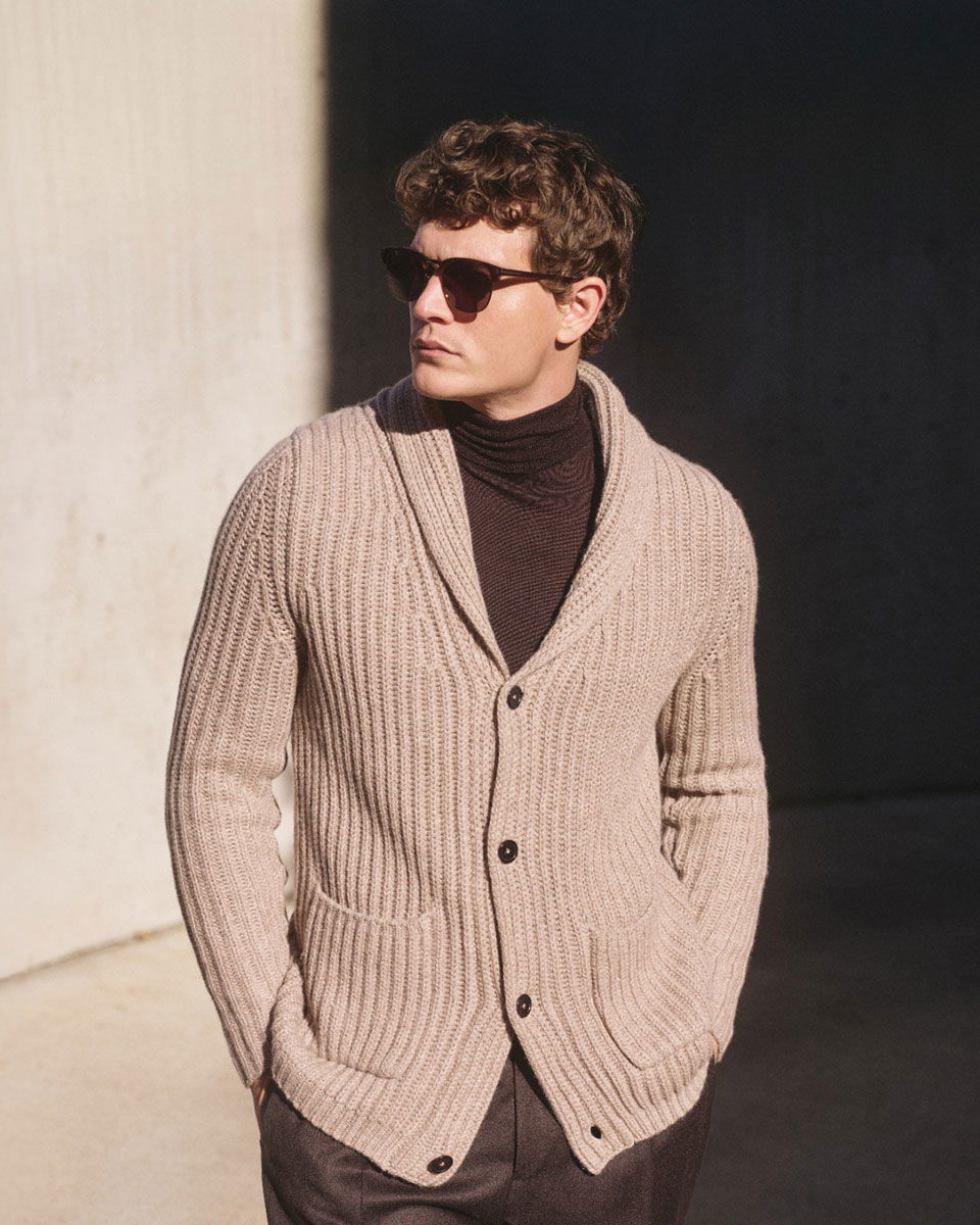 Man wearing dark sunglasses, a beige chunky knit cardigan with front pockets, and a dark turtleneck sweater, standing outdoors.