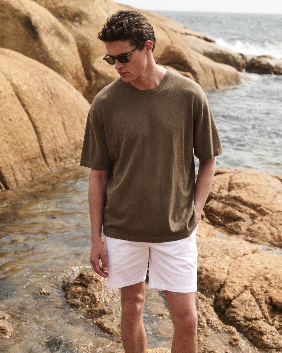 Man wearing sunglasses, an olive green short-sleeve shirt, and white shorts standing on rocky shore near the ocean.