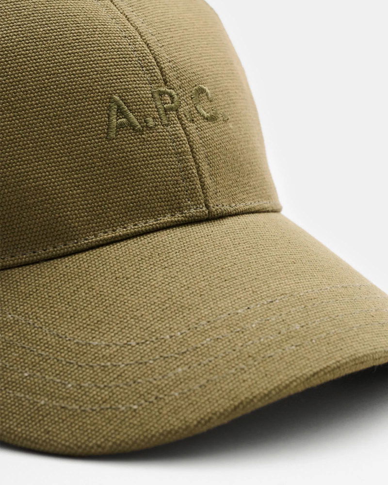 Close-up of an olive green textured fabric baseball cap with stitched 'A.P.C' logo on the front panel and curved brim.
