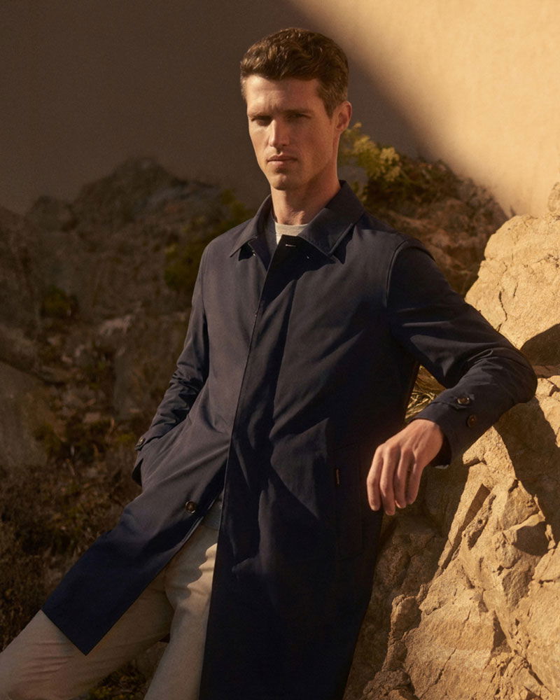 Mann lehnt entspannt an Felsen, trägt dunkelblauen Mantel und helle Hose, Hintergrund mit Felsen und warmem Licht.