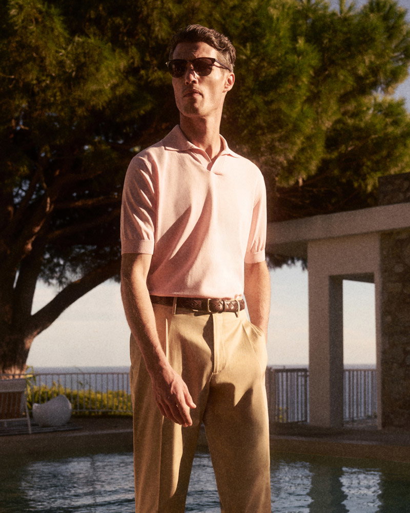 Man wearing sunglasses, a light pink short-sleeve polo shirt, and beige trousers standing by a pool with trees and a building in the background.
