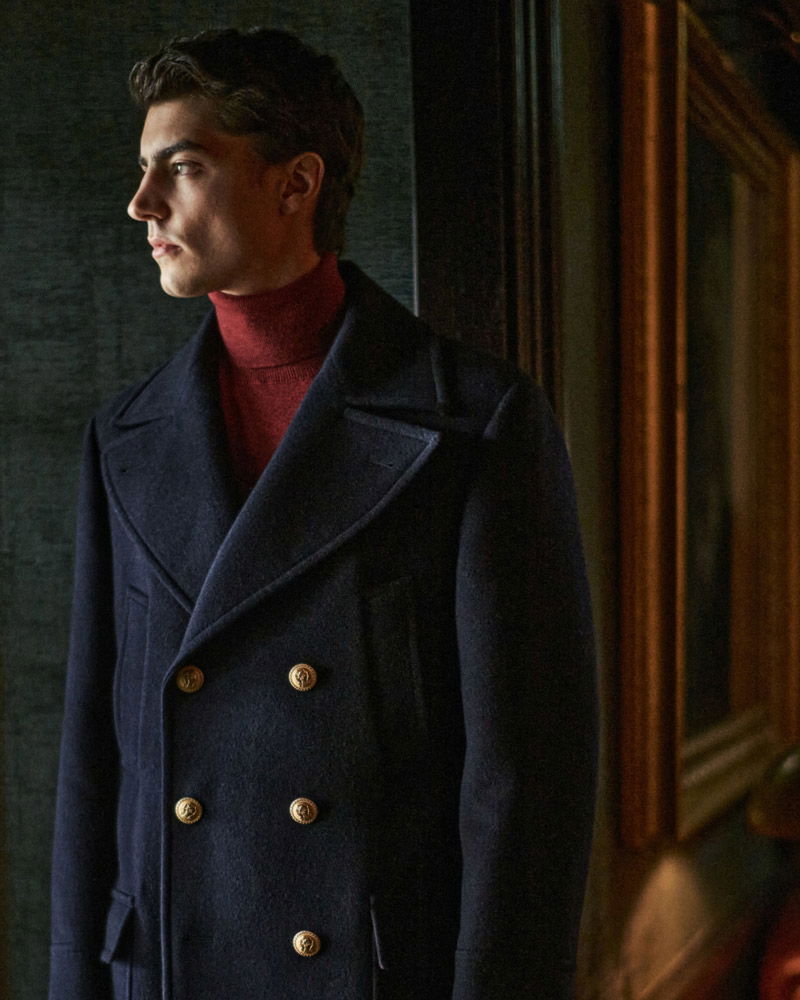 Young man with dark hair wearing a navy double-breasted coat and red turtleneck, looking to the left in dim light indoors.