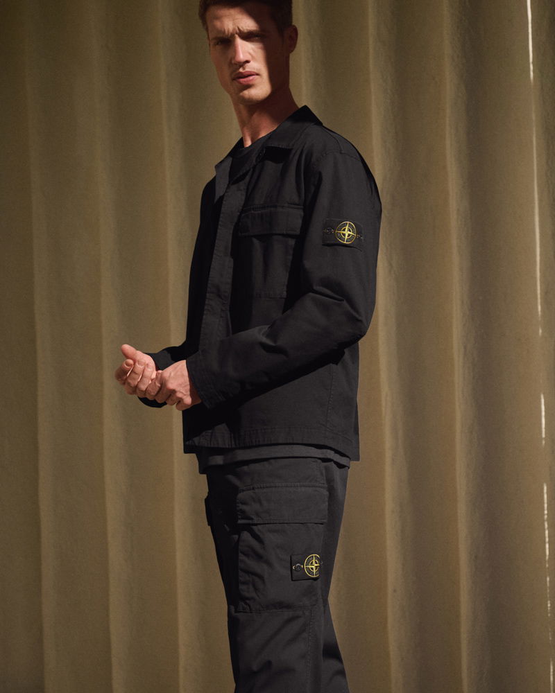 Mann trägt schwarze Stone Island Jacke und Cargo-Hose mit Logo-Patches an Ärmel und Bein vor beigem Vorhang.