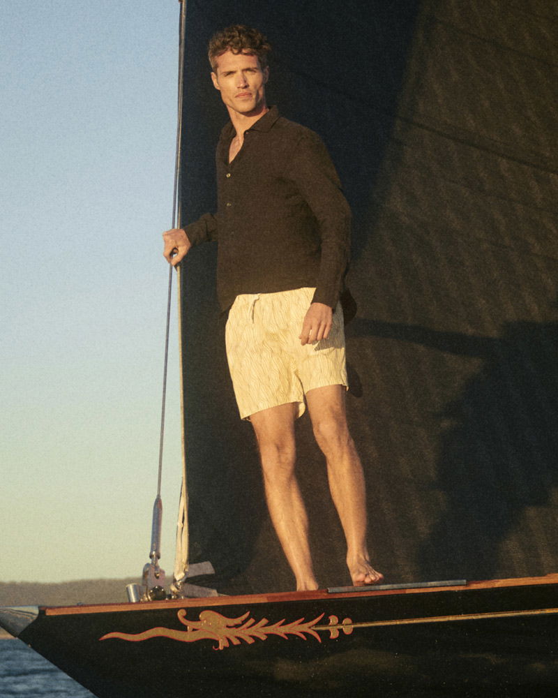 Mann mit dunklem Hemd und hellen Shorts steht barfuß an Bord eines Segelboots bei Sonnenuntergang.