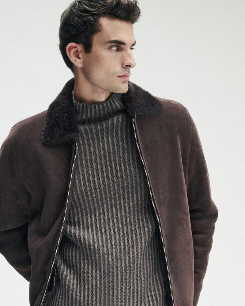 Junger Mann mit dunklen Haaren trägt braune Wildlederjacke mit Fellkragen und grauen, gestreiften Rollkragenpullover.