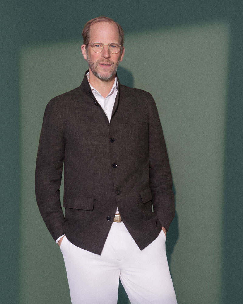 Mann mit Brille trägt braune Jacke, weiße Hose, steht vor grünem Hintergrund, Hände in den Taschen, schaut in die Kamera.