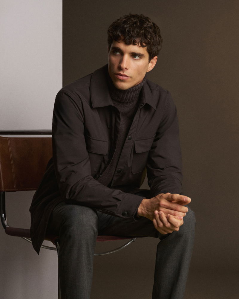 Young man with curly dark hair wearing a dark jacket and turtleneck, sitting thoughtfully on a brown chair against a two-tone background.