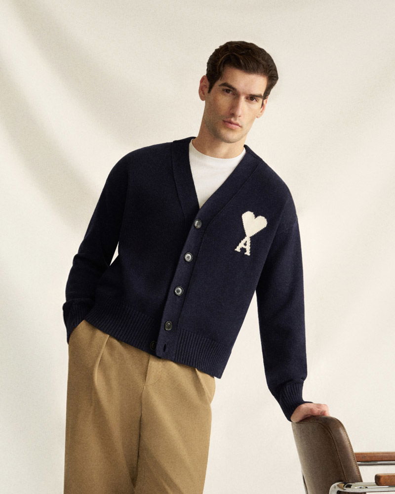 Mann trägt dunkelblaue Strickjacke mit Herz-Logo links und beige Hose, lehnt sich lässig an einen Stuhl.