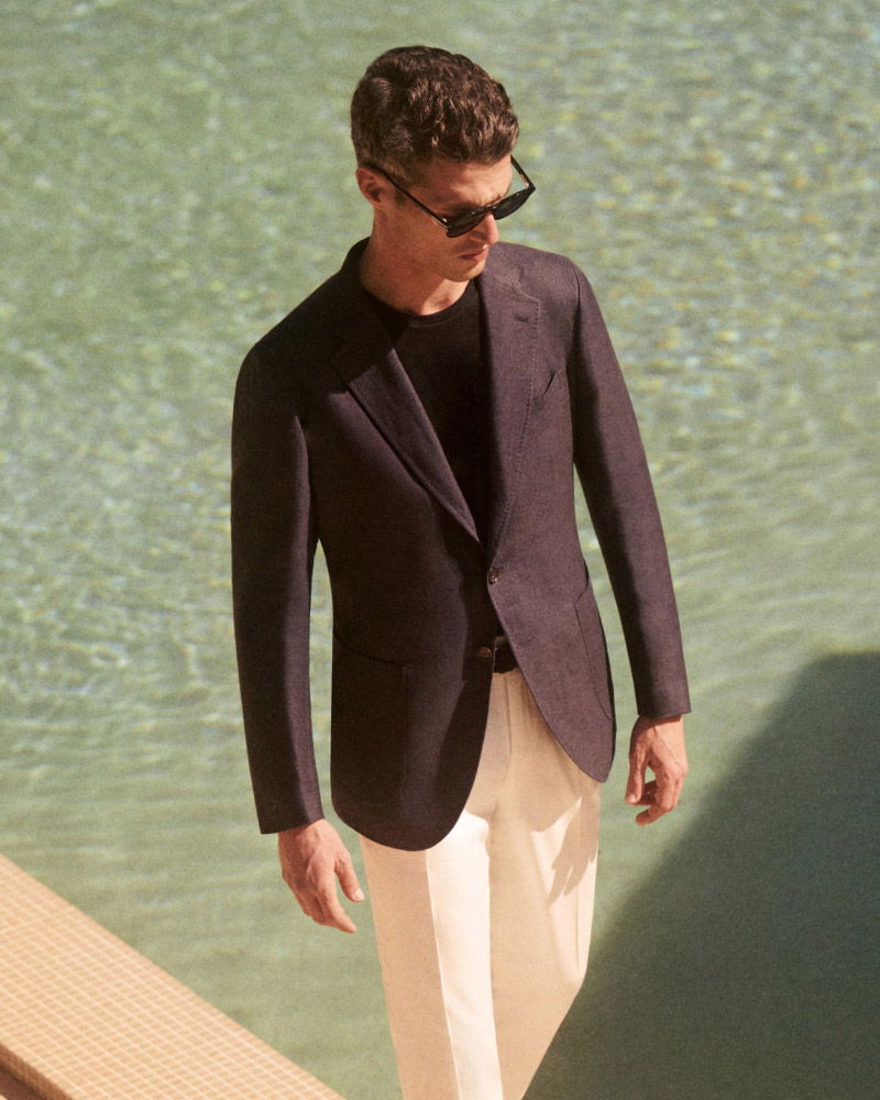 Mann mit dunkler Sonnenbrille trägt schwarzen Blazer, schwarzen Pullover und weiße Hose, steht am Beckenrand mit Wasser im Hintergrund.