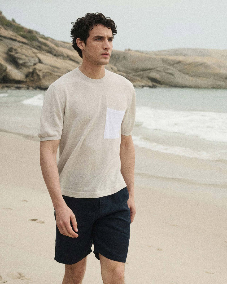 A man with curly hair wearing a beige short-sleeve shirt with a white pocket and black shorts walking on a sandy beach.