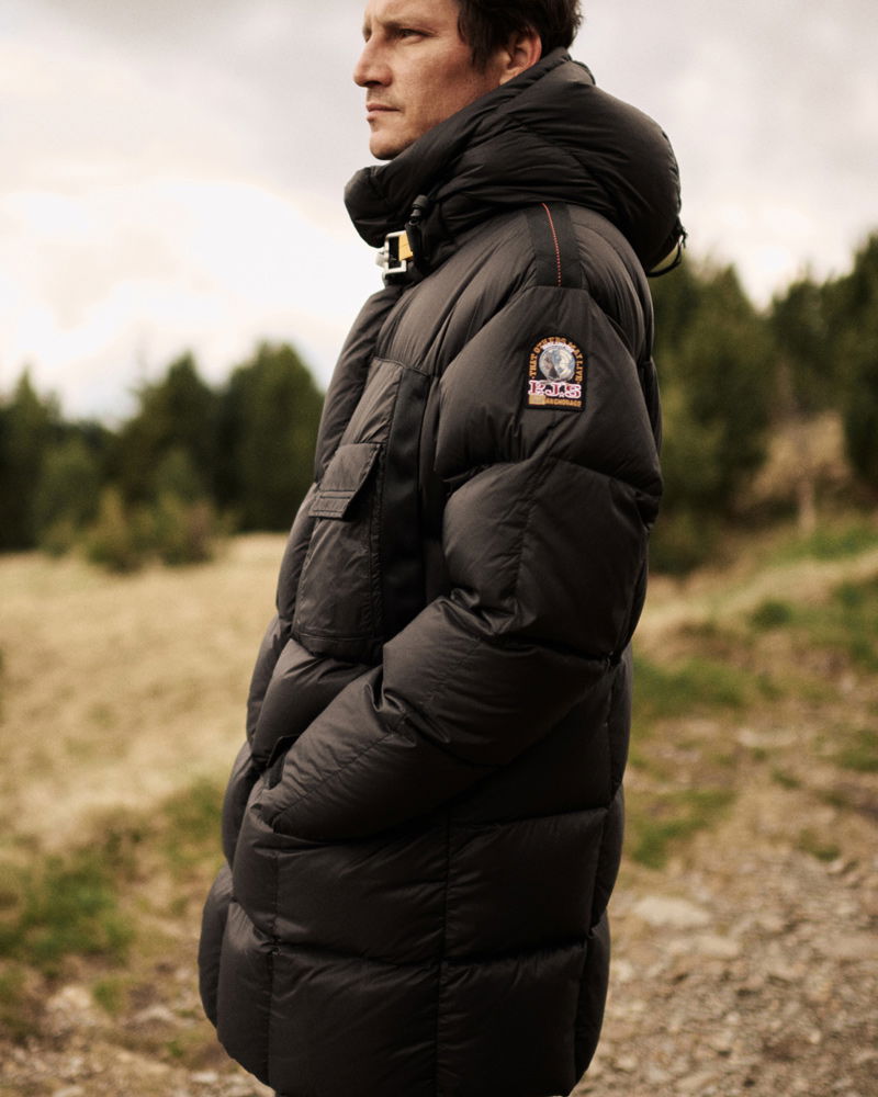 Side view of a man wearing a black puffer jacket with a hood, standing outdoors on a grassy path with trees in the background.