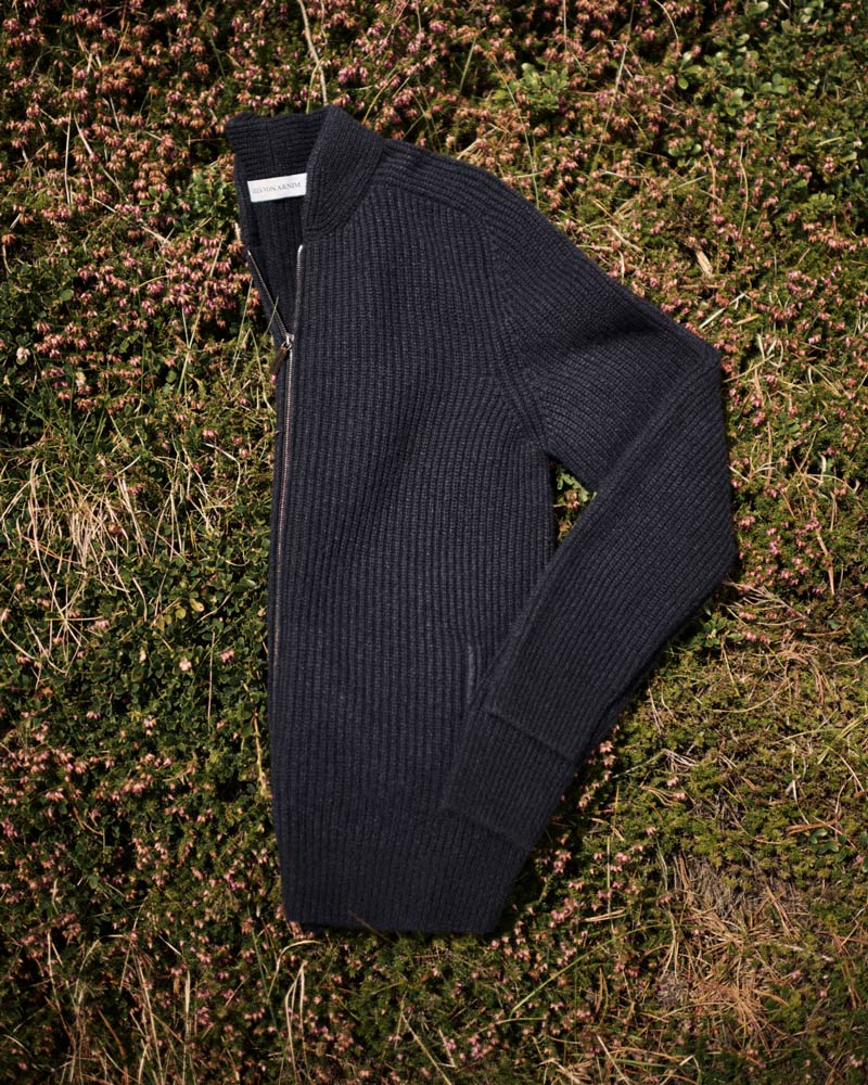Dunkelgrauer, grob gerippter Wollpullover mit Reißverschluss, liegend auf grünem, blühendem Heidekraut.