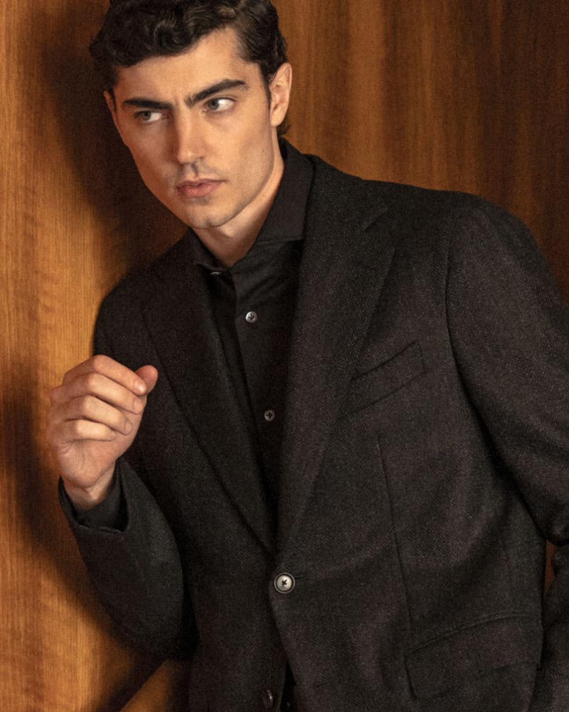 Junger Mann mit dunklem, gewelltem Haar trägt schwarzes Sakko und Hemd, lehnt vor braunem Holzhintergrund.