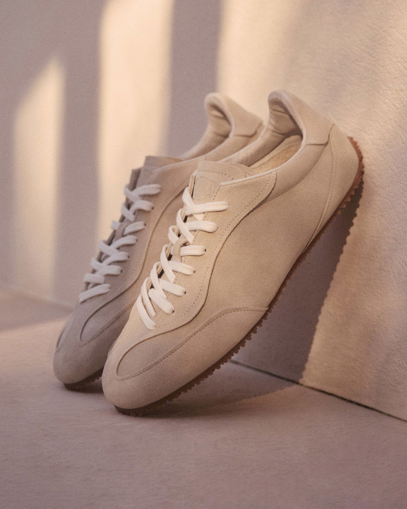 Beige Wildleder-Sneaker mit weißen Schnürsenkeln, brauner Profilsohle, stehend vor hellem Hintergrund und Schatten.
