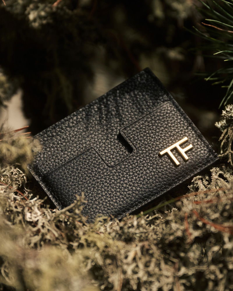 Black textured leather cardholder with gold 'TF' logo resting on natural moss and foliage background.