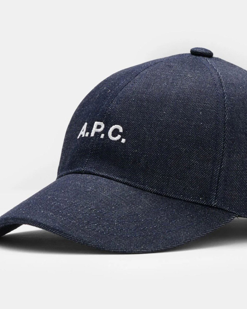 Dark blue denim baseball cap with white embroidered 'A.P.C.' logo on the front and a button on top, shown on a white background.