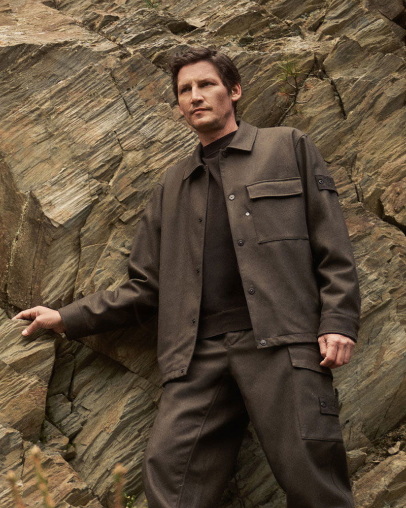 Man wearing dark brown jacket and pants standing against rugged rock formation outdoors, looking to the side with a serious expression.