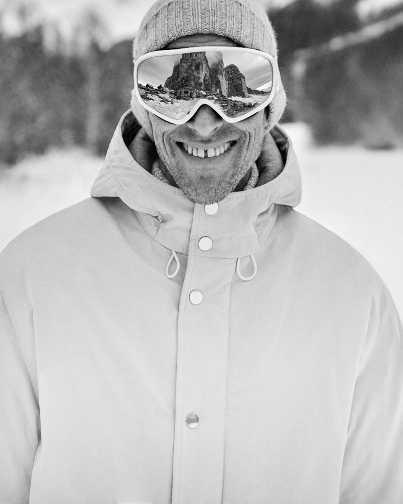 Mann im Wintermantel mit Mütze und Skibrille, die Berglandschaft mit Felsen reflektiert, lächelt in verschneiter Umgebung.