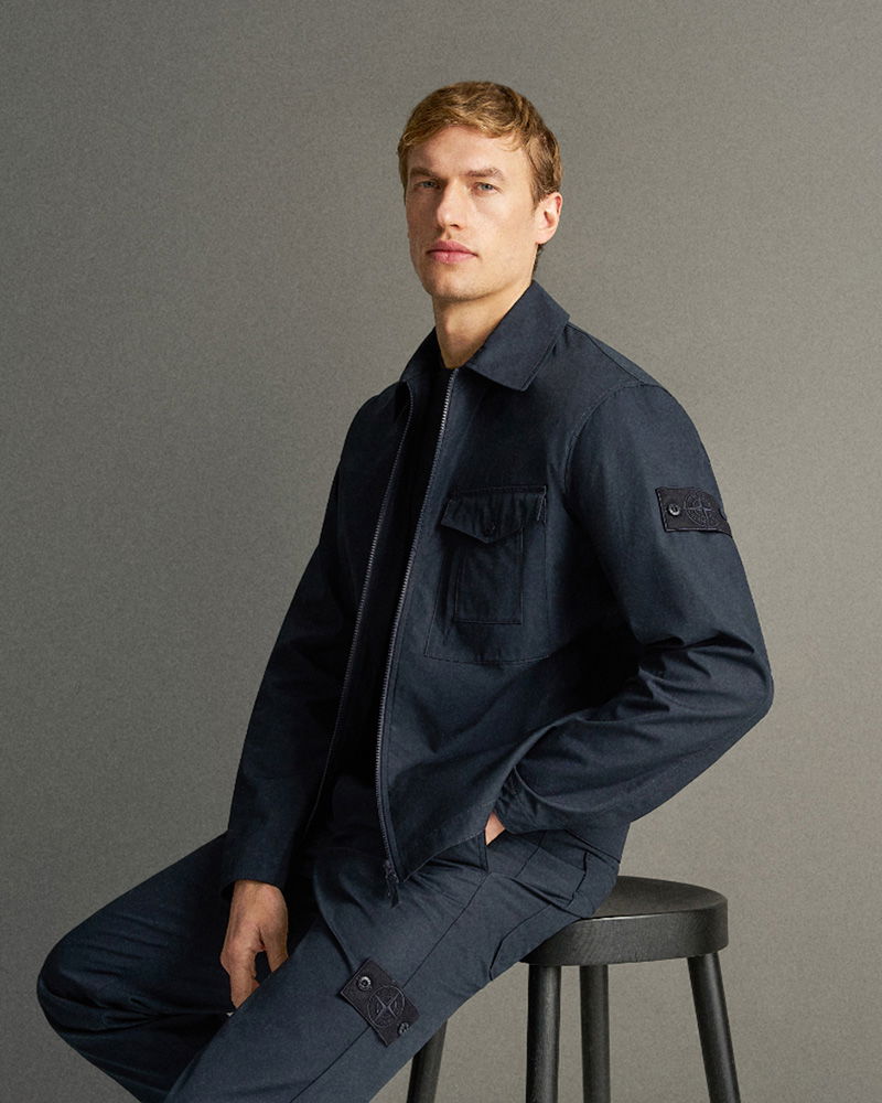 Mann mit blonden Haaren sitzt auf Hocker, trägt dunkle Jacke und Hose mit aufgesetzten Taschen und Emblem am Ärmel.