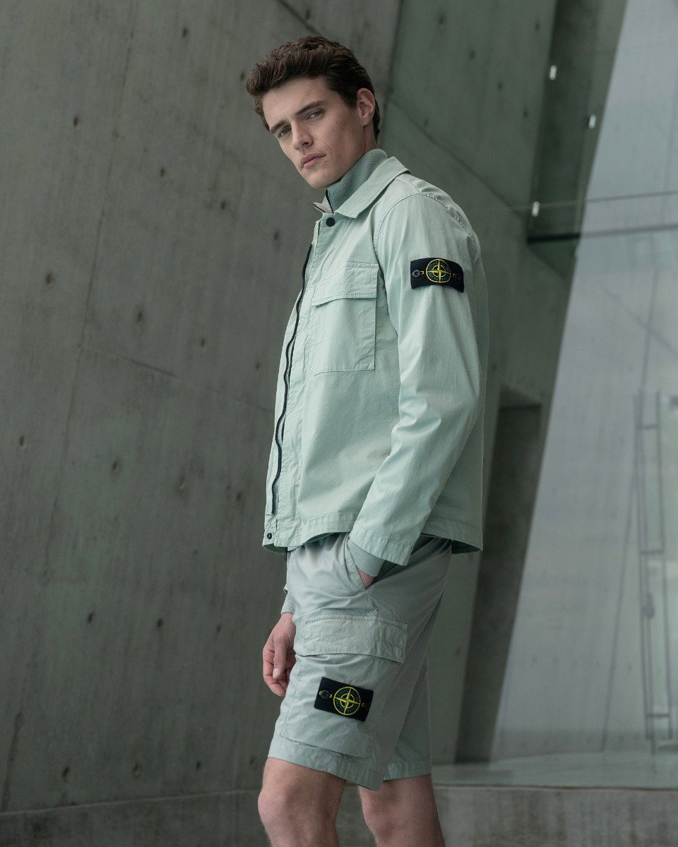Young man wearing light green jacket and matching shorts with black and yellow logo patches, standing against a concrete wall.