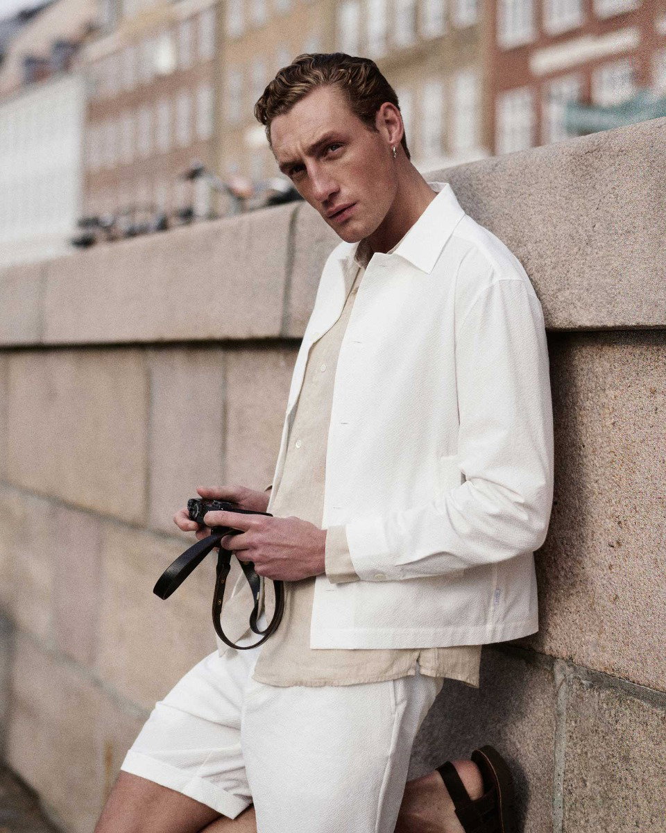 Young man with short curly hair wearing a white jacket, beige shirt, and white shorts, holding a camera while leaning against a stone wall.