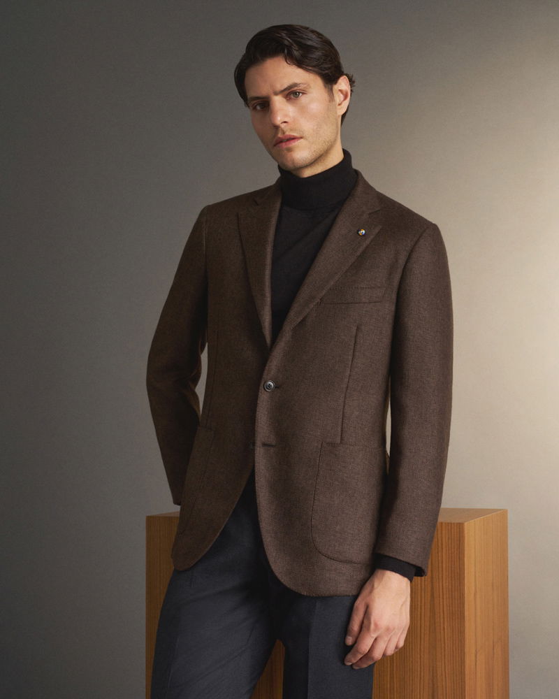 Man wearing a brown blazer over a black turtleneck, standing next to a wooden pedestal against a gradient beige-gray background.