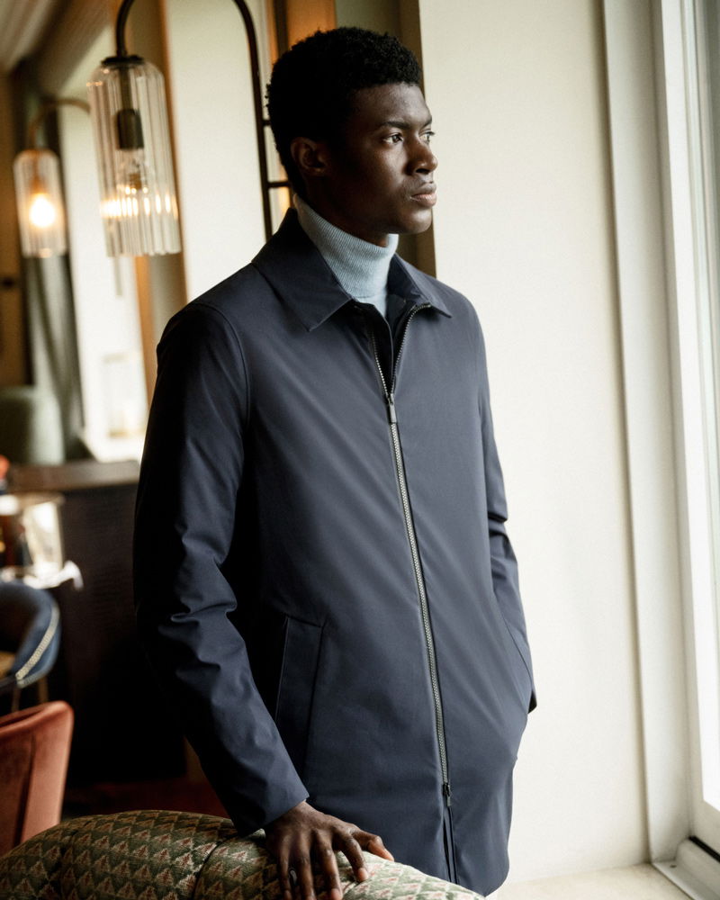 Mann mit dunklem Mantel und grauem Rollkragenpullover steht nachdenklich am Fenster in einem stilvollen, warm beleuchteten Raum.