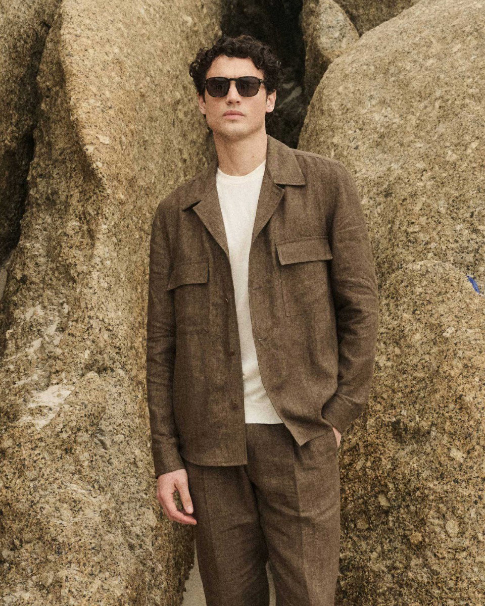 Man wearing brown linen jacket and pants with a white shirt and black sunglasses standing against large textured rocks.
