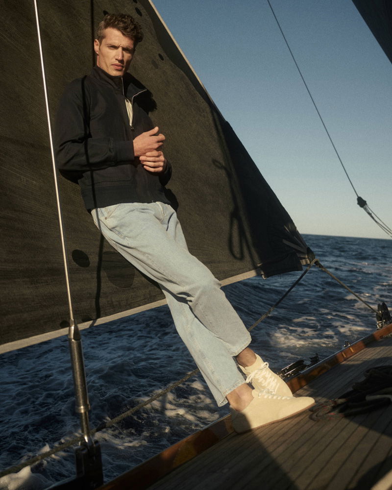 Junger Mann in schwarzer Jacke, hellen Jeans und weißen Schuhen lehnt an schwarzem Segel auf Segelboot bei sonnigem Wetter.