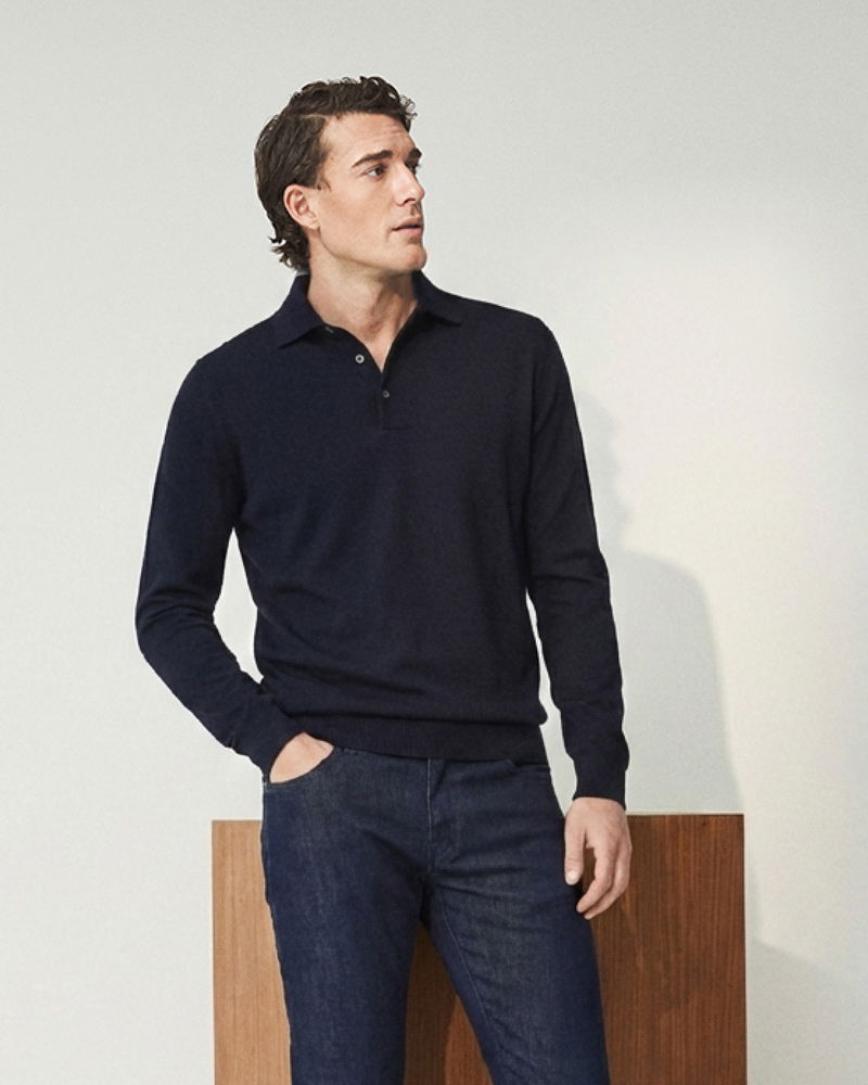 Mann mit dunklem Poloshirt und Jeans steht vor hellem Hintergrund, eine Hand in Hosentasche, schaut seitlich nach oben.