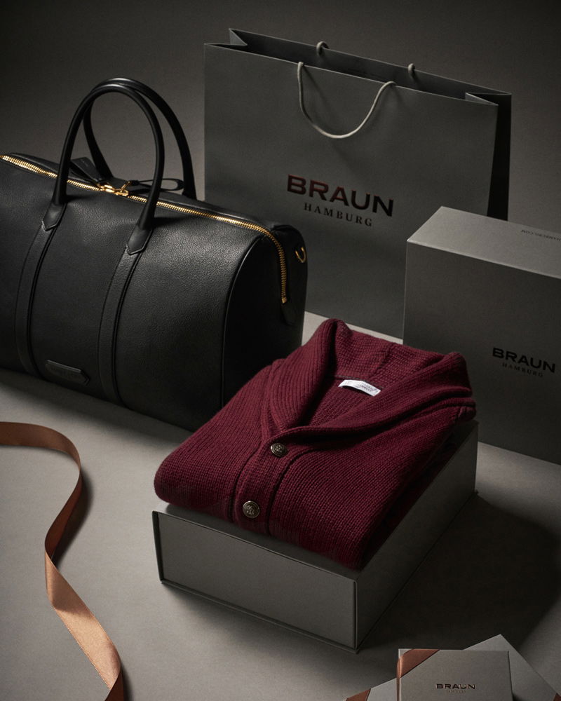Schwarze Lederreisetasche, graue Geschenktüten und -boxen mit Braun Hamburg Logo, rote Strickjacke und braunes Geschenkband.