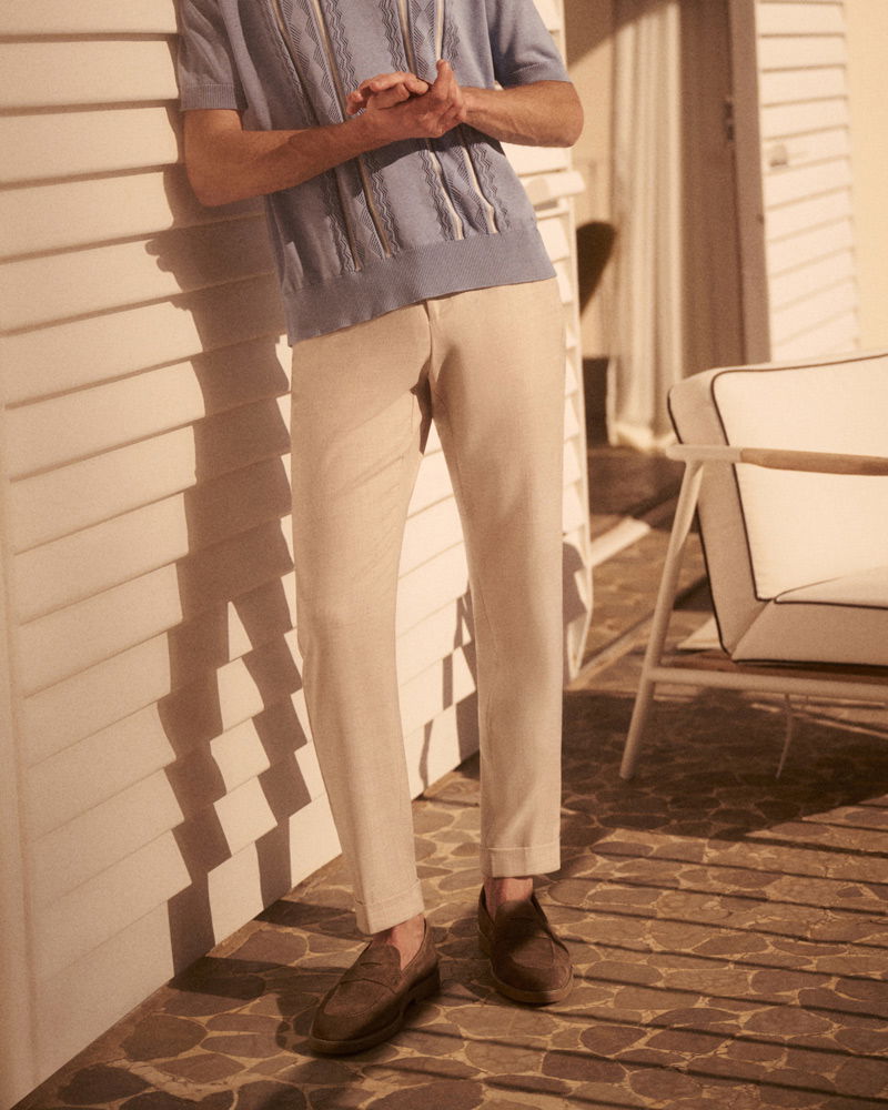 Mann lehnt an weißer Wand, trägt hellblaue Strickjacke, beige Hose und braune Loafers, neben weißem Gartenstuhl.