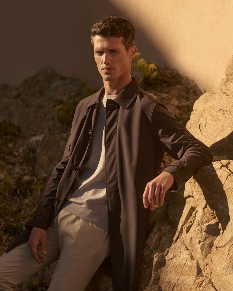 Mann lehnt an Felsen, trägt beigen Mantel, graues Shirt und helle Hose, Sonnenlicht wirft Schatten auf Gesicht und Felsen.