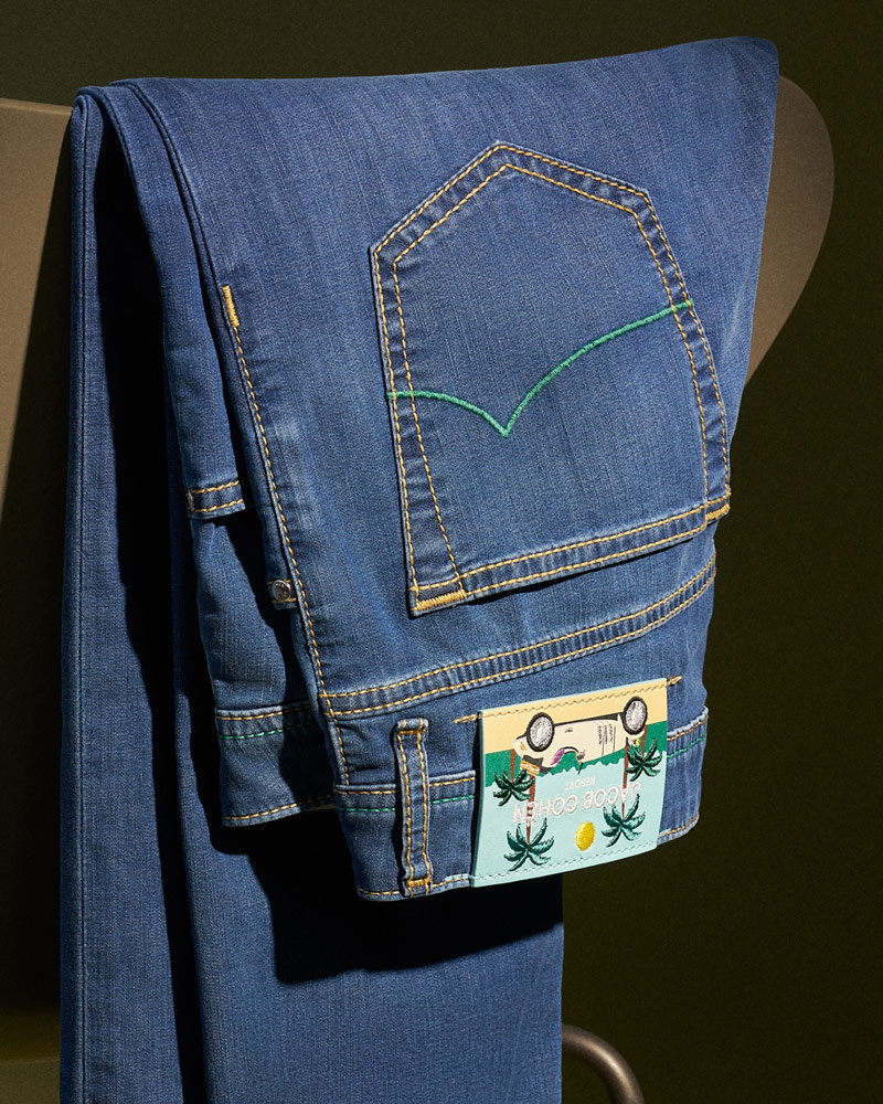 Folded blue denim jeans with yellow and green stitching, displayed hanging over a dark brown chair against a dark background.