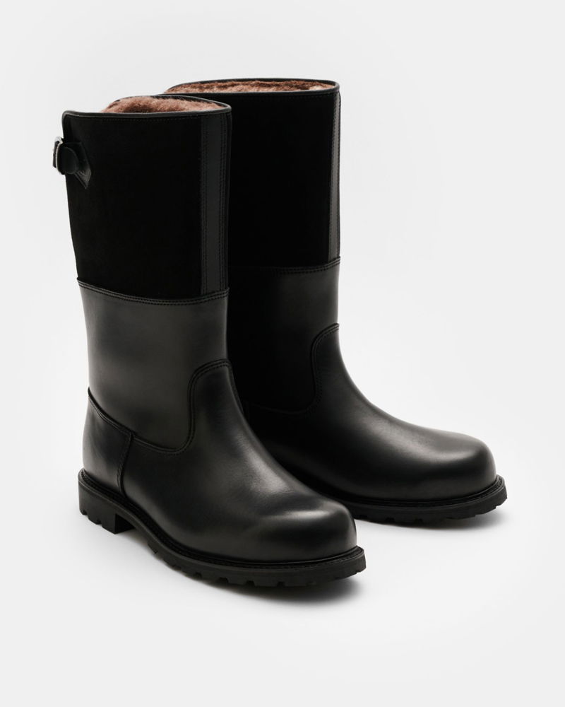 Pair of black mid-calf leather boots with suede upper section, brown fur lining, buckle detail, and rugged sole on white background.
