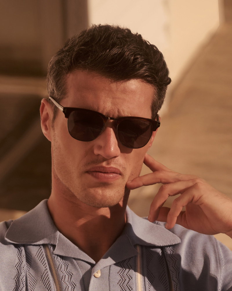 Close-up of a man wearing dark sunglasses and a light blue patterned shirt, touching his face with a thoughtful expression.