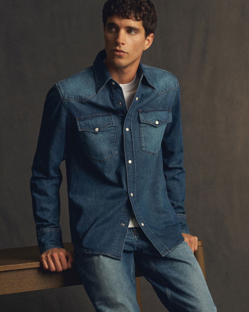Young man with curly hair wearing a buttoned denim shirt over a white t-shirt and blue jeans, sitting on a wooden table.