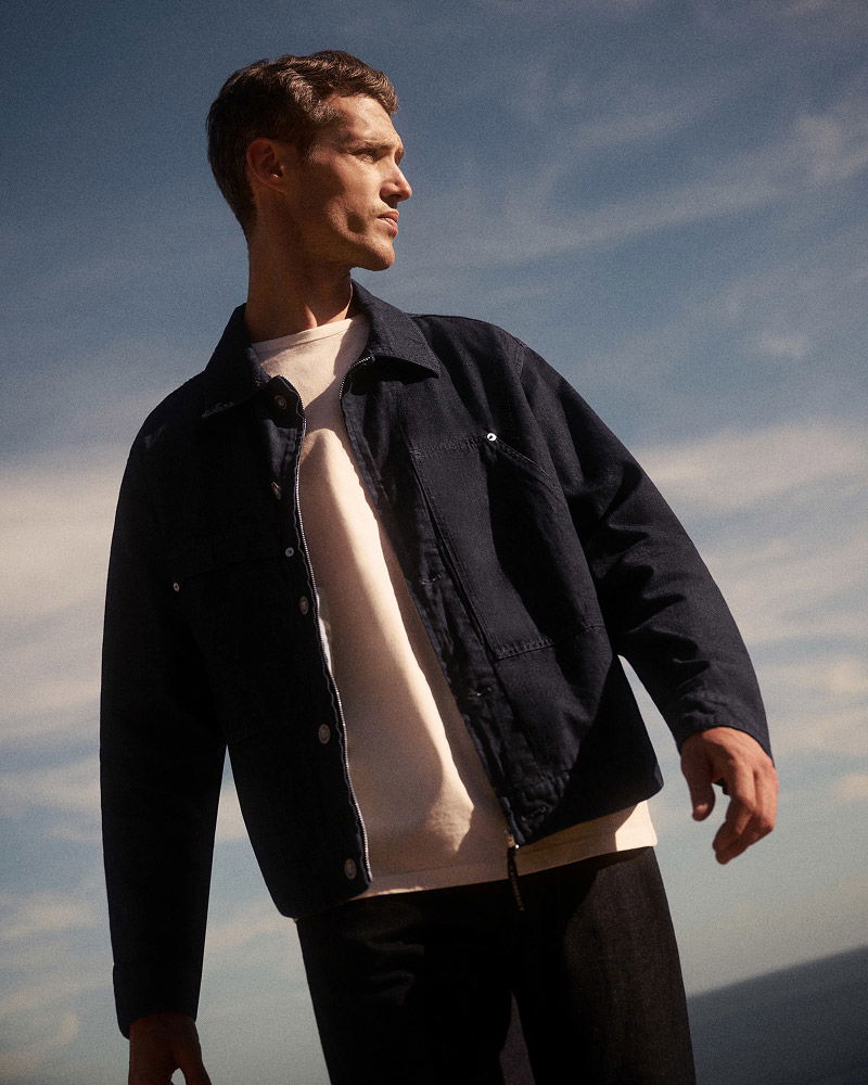 Junger Mann mit kurzem Haar trägt weiße Shirt und offene dunkelblaue Jacke, blickt nach links vor bewölktem Himmel.