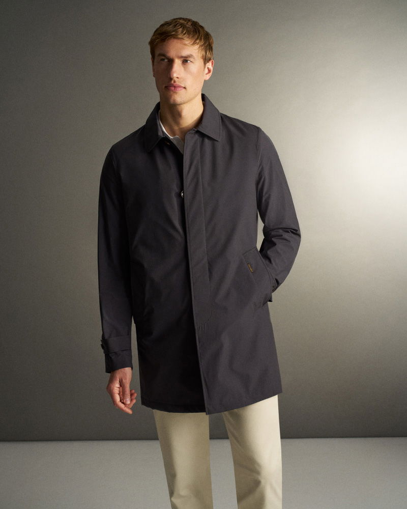 Mann mit blondem Haar trägt dunkelblauen Mantel und helle Hose, steht vor neutralem grauem Hintergrund, rechte Hand in Manteltasche.