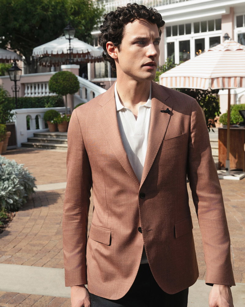 Junger Mann mit lockigem Haar trägt braunes Sakko und weißes Poloshirt, steht vor Terrasse mit Pflanzen und Sonnenschirmen.