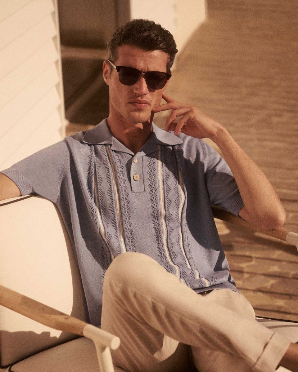 Man wearing dark sunglasses, light blue patterned short-sleeve polo shirt, and beige pants, sitting on a chair outdoors.