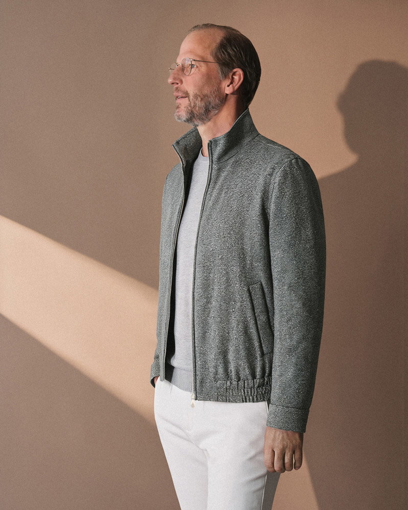 Middle-aged man with glasses wearing a gray zip-up jacket and white pants standing against a beige wall with sunlight and shadow.