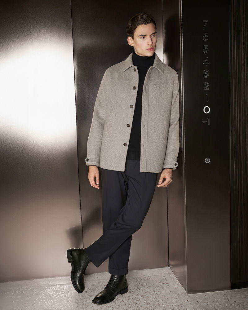 Young man wearing a beige jacket, black turtleneck, navy pants, and black boots, standing in an elevator by floor buttons.