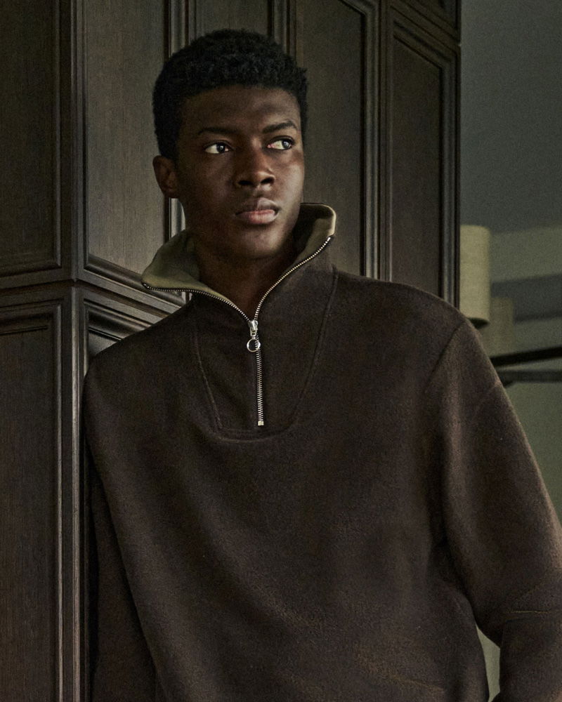 Young man wearing a dark brown half-zip pullover sweater standing indoors against wooden paneling, looking to the side.
