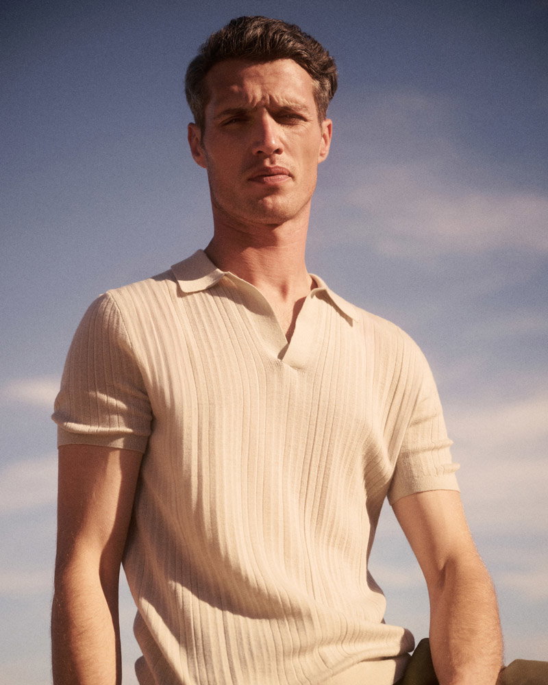Young man wearing a beige ribbed short-sleeve polo shirt standing outdoors under a partly cloudy sky.